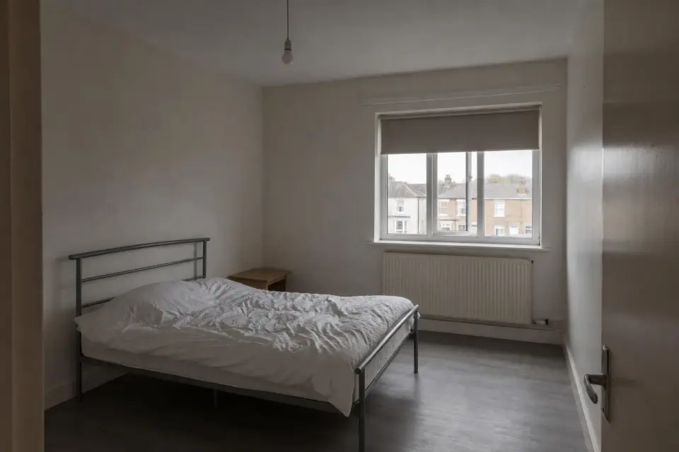 A bare rental bedroom with off-white walls, a metal bed frame and a roller blind.